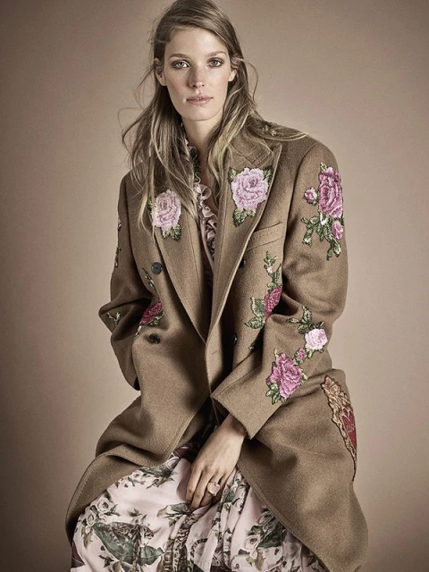 A blonde adult female poses in a long, floral dress with a brown, embroidered overcoat in a studio environment with soft lighting.