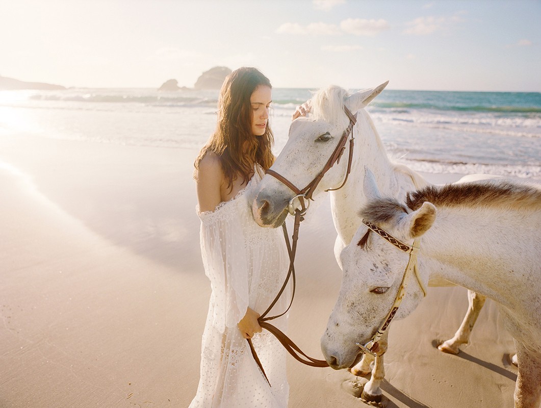 A woman in a white dress stands with two white horses on a sandy beach with gentle waves, under a bright sky with a soft, golden light.