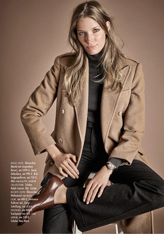 A fashionable adult female poses in a tan double-breasted wool coat, black turtleneck, and brown leather loafers in a studio shot.