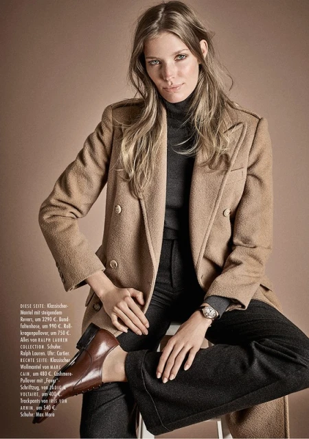 A fashionable adult female poses in a tan double-breasted wool coat, black turtleneck, and brown leather loafers in a studio shot.