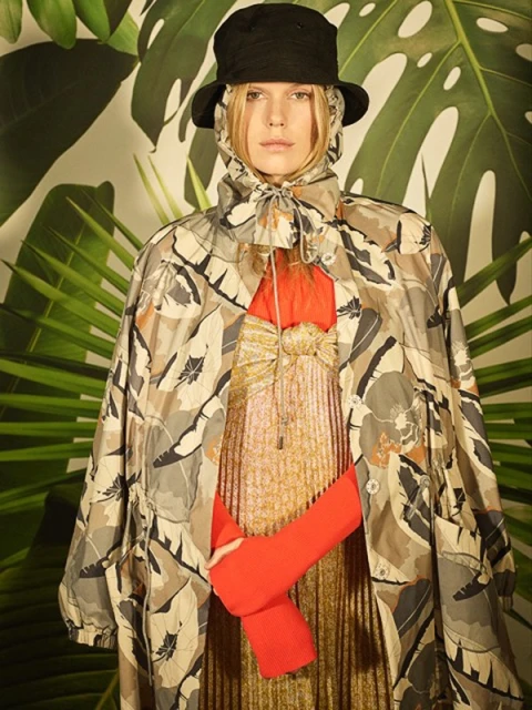 Blonde model poses in a leaf-patterned poncho, orange top, and a black hat against a backdrop of tropical palm fronds.