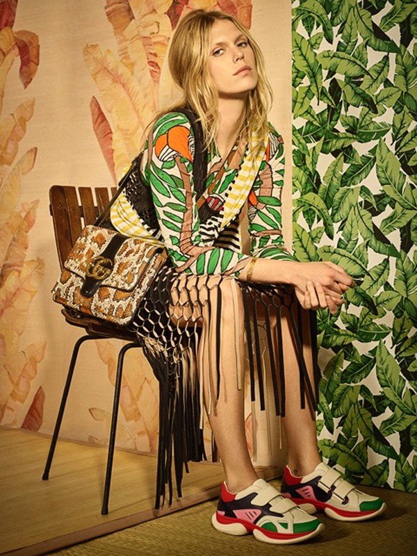 Fashionable blonde model sits in a photo studio wearing a printed dress, fringed vest, platform sneakers, and a Gucci handbag with jungle-themed wallpaper.