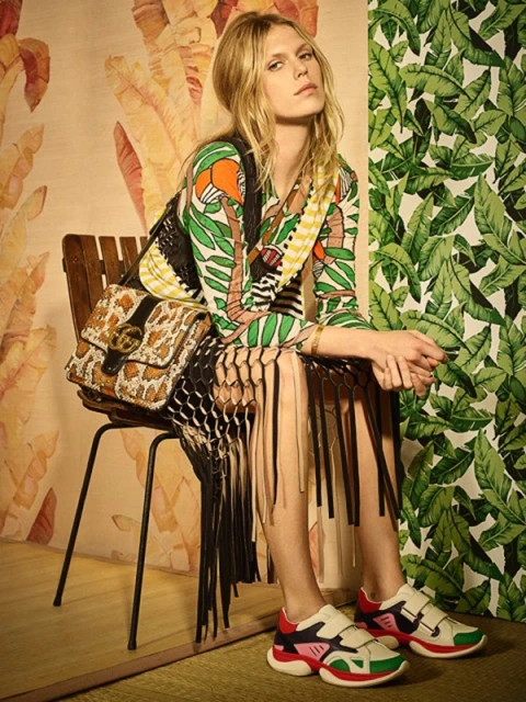Fashionable blonde model sits in a photo studio wearing a printed dress, fringed vest, platform sneakers, and a Gucci handbag with jungle-themed wallpaper.