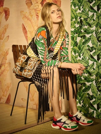 Fashionable blonde model sits in a photo studio wearing a printed dress, fringed vest, platform sneakers, and a Gucci handbag with jungle-themed wallpaper.