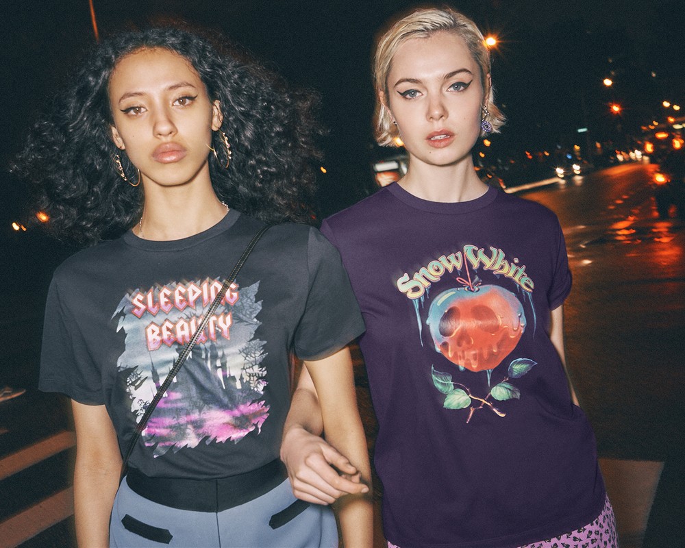 Two women pose closely together at night, showcasing graphic t-shirts with city lights blurred in the background.