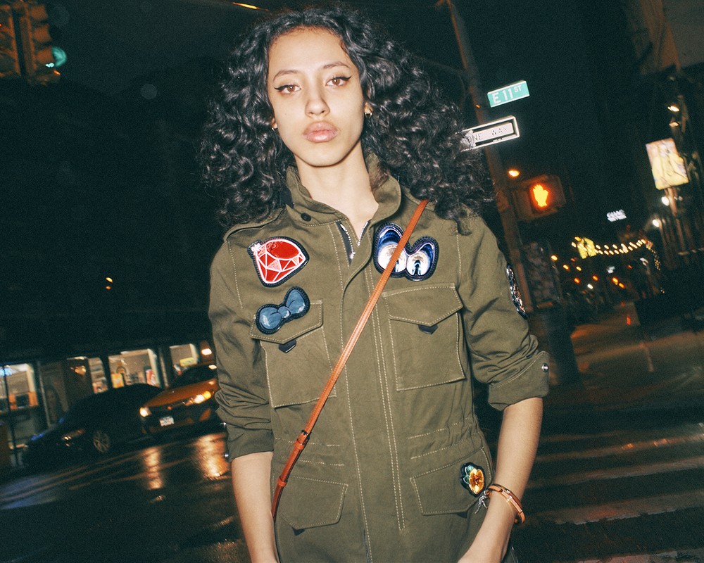 Young woman in urban setting at night. She has dark curly hair and is wearing a military-style jacket with patches. City lights visible.