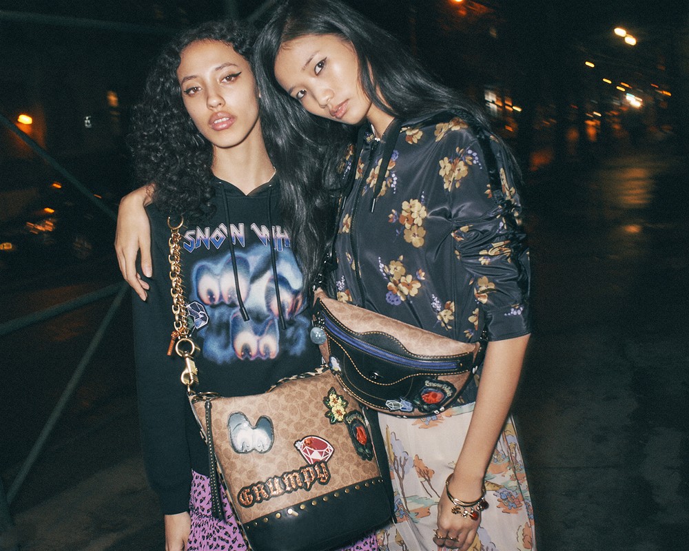 Two young women pose outdoors at night with designer bags. One wears a floral jacket, and the other, a Snow White hoodie.