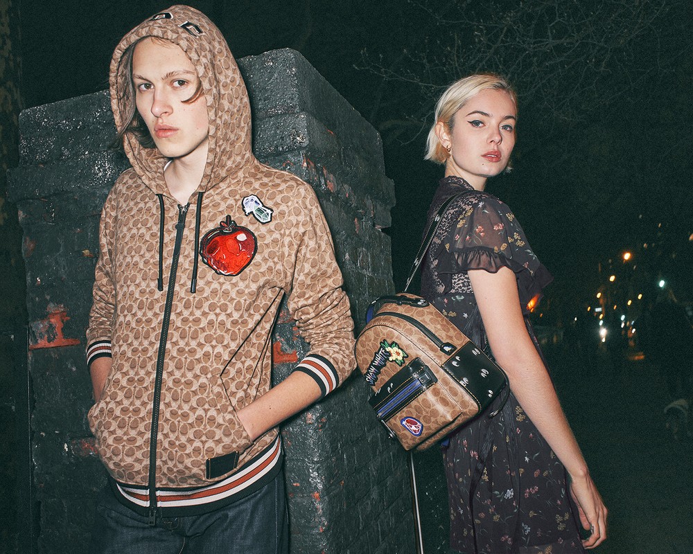Two people, a man and a woman, stand outdoors at night with dim lighting. The man wears a patterned hoodie and the woman a floral dress.