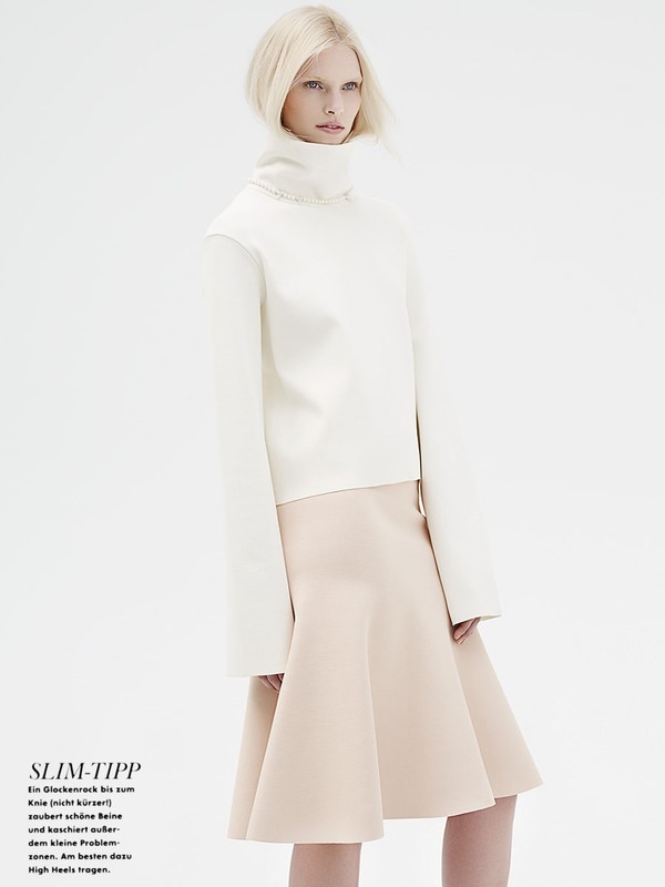 Model in white turtleneck sweater and beige skirt against a plain white seamless studio background.