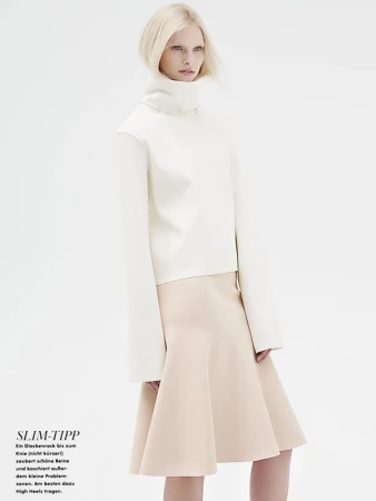 Model in white turtleneck sweater and beige skirt against a plain white seamless studio background.