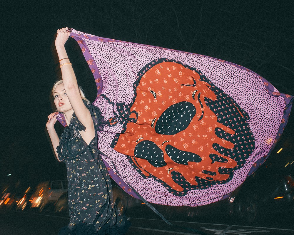 Blonde woman holding a large printed scarf with a skull design. Dark background with blurred vehicle lights at night.