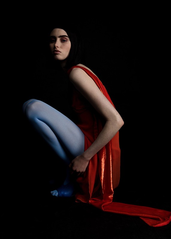 Woman in red dress and blue tights squats in dark studio. Intense gaze, dramatic lighting, and contrasting colors create striking fashion image.