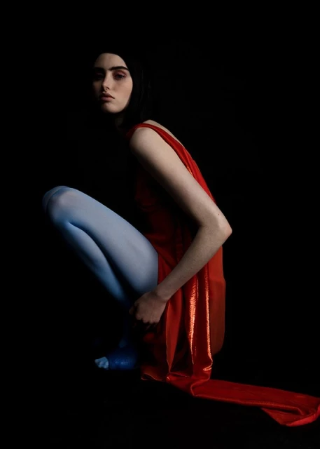 Woman in red dress and blue tights squats in dark studio. Intense gaze, dramatic lighting, and contrasting colors create striking fashion image.