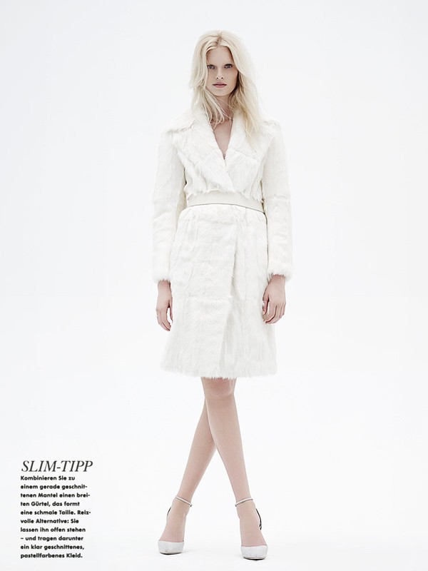 Fashion model in a full-length pose wearing a white fur coat and matching heeled shoes in a bright, neutral studio setting.