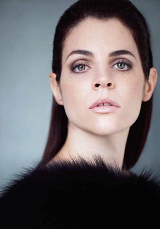 Close-up studio portrait of a fair-skinned adult woman with dark hair, wearing black furry top, against a light blue background.