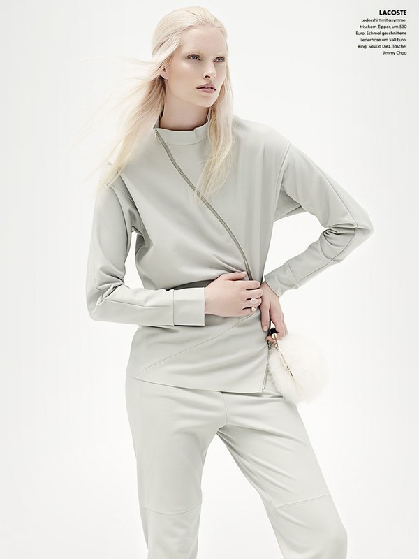 A fair-skinned woman poses in a pale gray outfit, holding a white fur purse against a bright white background, as part of a fashion editorial.