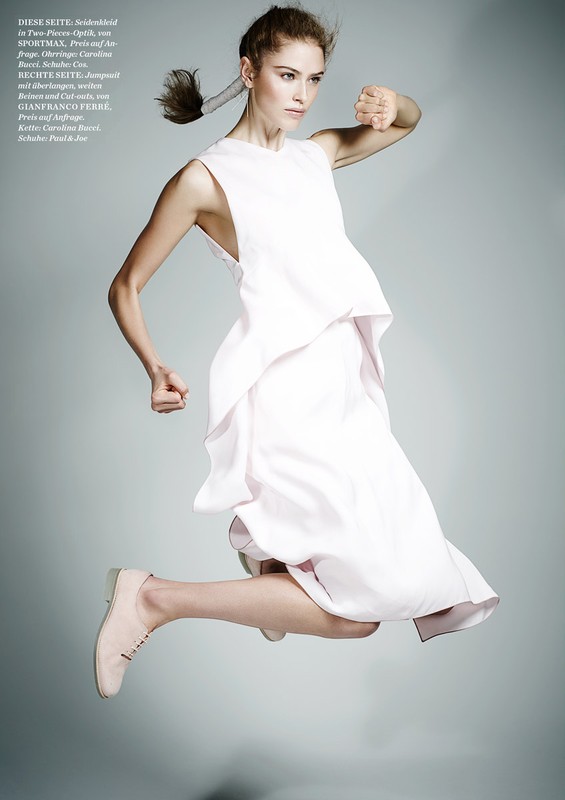 A woman in a flowing white dress jumps mid-air against a pale gray backdrop, showcasing an energetic and stylish fashion pose.