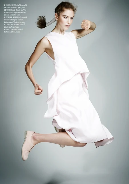 A woman in a flowing white dress jumps mid-air against a pale gray backdrop, showcasing an energetic and stylish fashion pose.