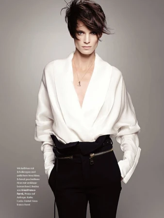 Model with short dark hair wearing a white silk blouse and black trousers against a neutral grey studio background.