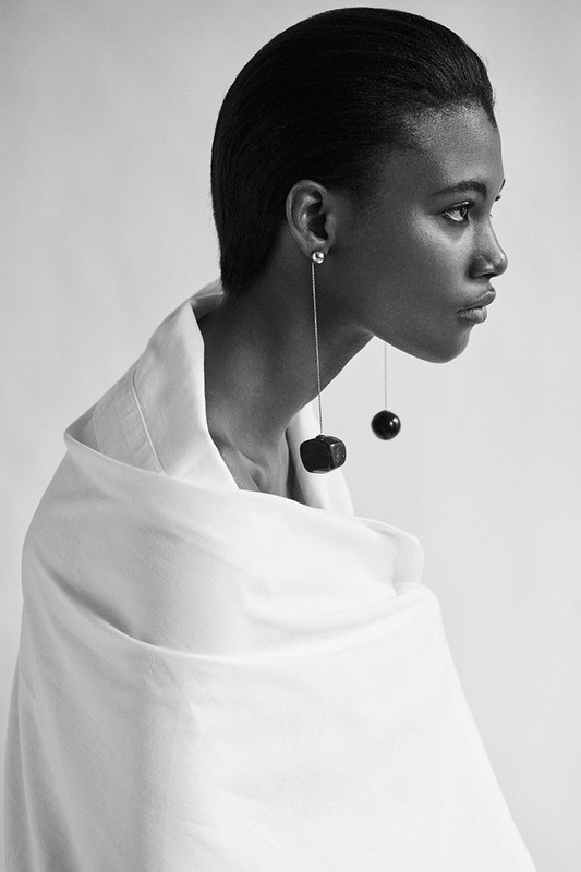 Black and white portrait of a Black woman in profile wearing statement earrings and a white collared shirt or shawl, with a neutral backdrop.