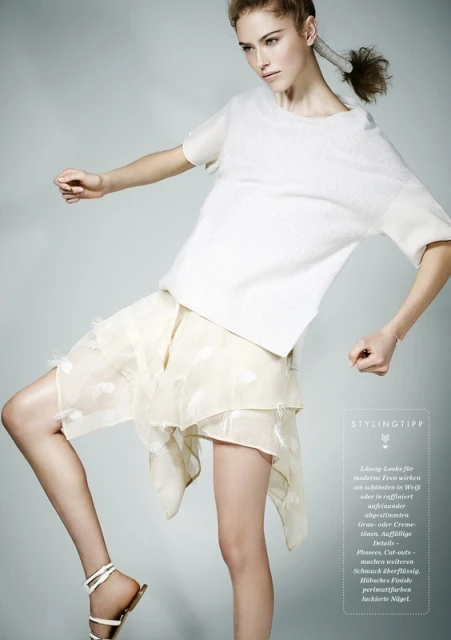 A young adult fashion model poses in a white top and layered cream skirt with strappy sandals in a studio fashion shot.
