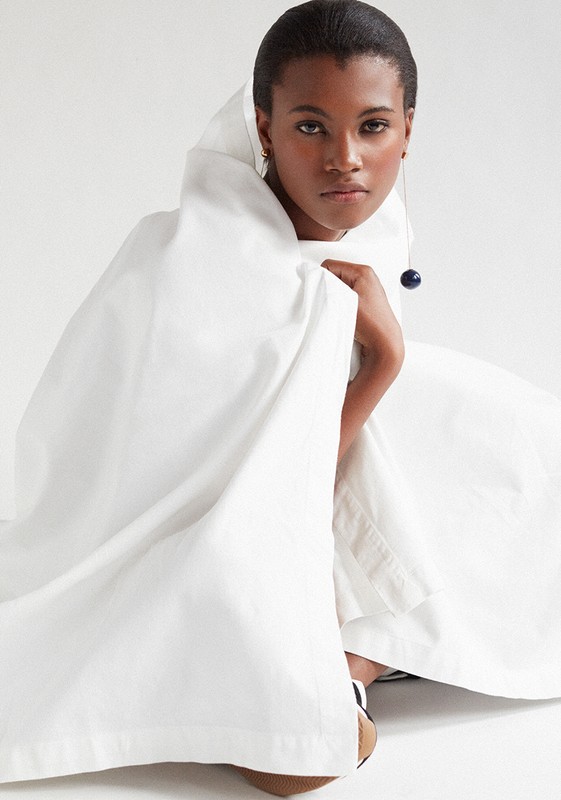 A single African-American adult female model wearing a white dress with a hood, posing in a photo studio with neutral lighting.