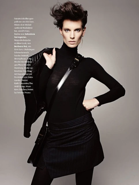 Fashionable woman in black turtleneck, mini skirt, and jacket poses in a studio, showcasing edgy and stylish look with dark makeup.