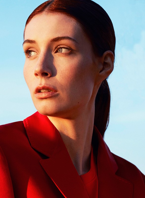 Headshot of a redhead woman wearing a red blazer looking off-camera in a bright, outdoor setting with a clear, pale blue sky background.