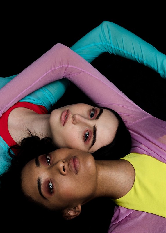 Two women lie head-to-head in colorful outfits against a black backdrop, creating a striking image with contrasting hues and tones.