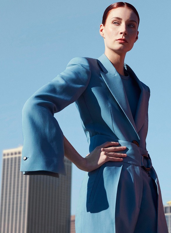 Adult woman in a blue pantsuit posing against a blue sky and urban skyline in a fashion editorial outdoor portrait.