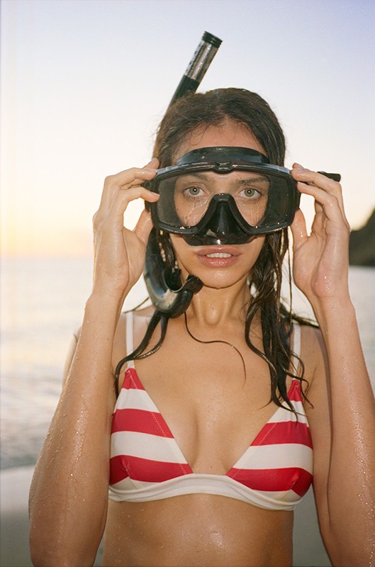 Woman in striped bikini wearing snorkel gear on sandy beach with ocean and distant land mass in soft, warm light at sunset or sunrise.