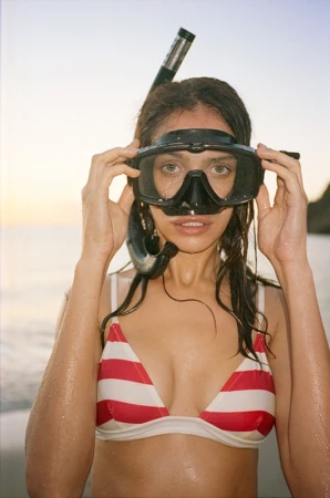 Woman in striped bikini wearing snorkel gear on sandy beach with ocean and distant land mass in soft, warm light at sunset or sunrise.