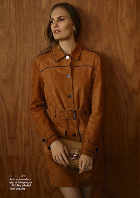 Fashion editorial shot of a woman in a brown suede dress with matching purse, posing against a wood panel backdrop in soft studio light.