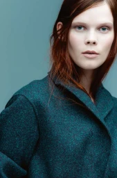Portrait of a red-haired woman wearing a textured dark green coat against a muted teal background.