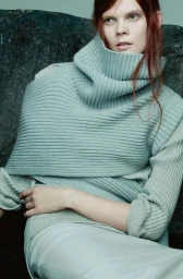 Model with red hair wearing a sage green turtleneck sweater posed against a dark textured stone background