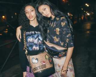 Two young female models posing outdoors at night wearing printed hoodies and designer crossbody bags with patches