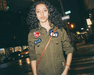 Young woman with dark curly hair wearing an olive green utility jacket decorated with patches on a city street at night.