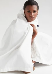 Black woman wearing draped white garment with drop earrings against plain background