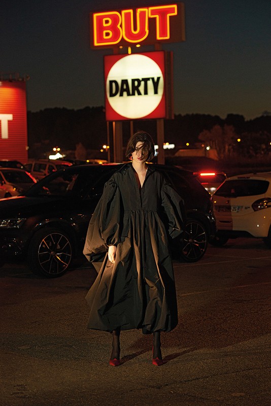 Woman in a black dress stands in a parking lot at night with 'BUT' and 'DARTY' signs behind her under the dark sky.