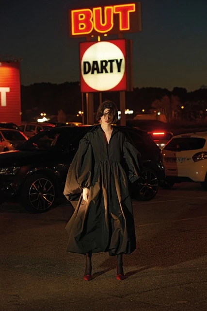 Woman in a black dress stands in a parking lot at night with 'BUT' and 'DARTY' signs behind her under the dark sky.