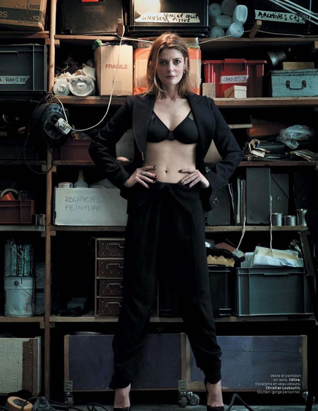 A woman in a black bra, blazer, and pants stands in front of a cluttered storage shelf with boxes, cans, and various stored items.