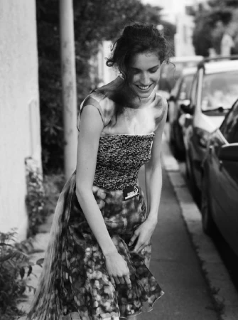 Black and white portrait of a smiling woman standing on a city sidewalk next to parked cars, photographed by Sarra Na in Los Angeles.