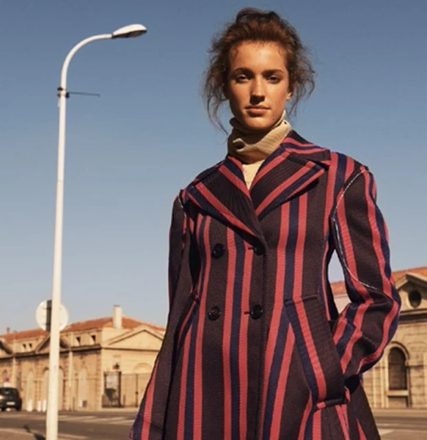 Woman in a striped coat stands in front of a building and blue sky. The light post adds a touch of urban elegance to the scene.