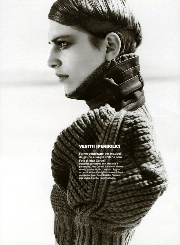 Black and white fashion portrait of a slender adult woman in a thick knit sweater and leather collar in a bright, overcast outdoor setting.