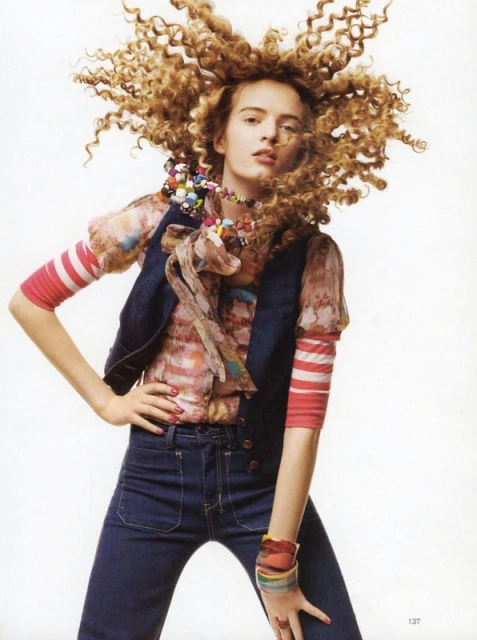 A woman with voluminous, curly blonde hair poses in a sleeveless denim vest, striped sleeves, jeans, and a beaded necklace against a white background.