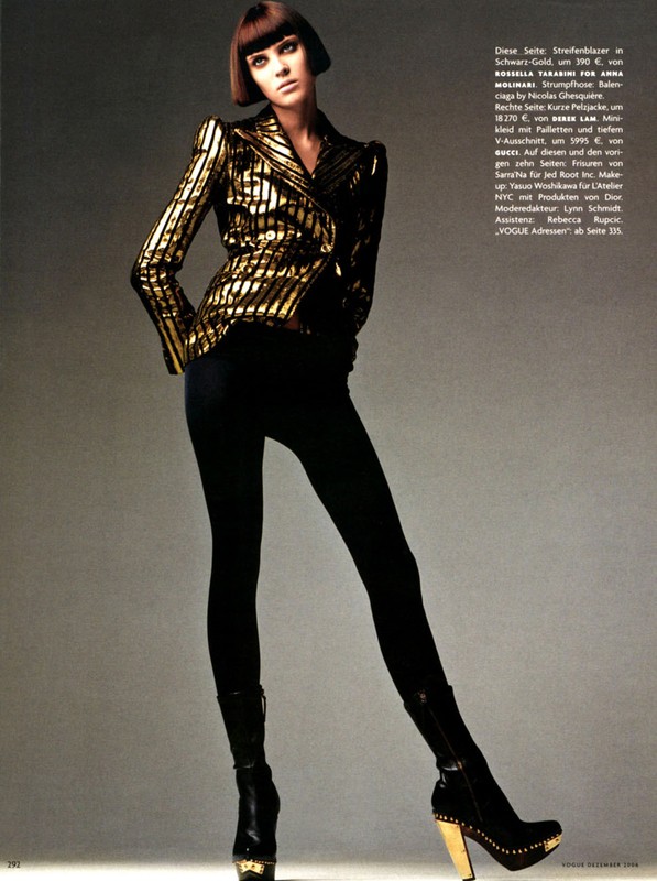 A woman wearing a metallic gold and black striped blazer and black leggings poses against a grey studio backdrop.