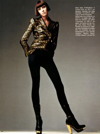 A woman wearing a metallic gold and black striped blazer and black leggings poses against a grey studio backdrop.