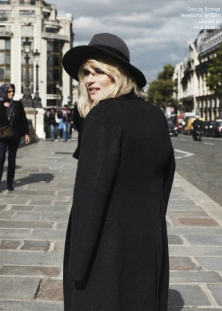 Blonde woman in black coat and dark fedora hat looking back on an urban street.