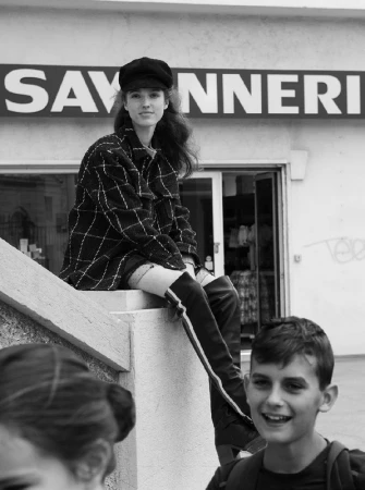 Monochrome portrait of a young woman seated on a stone ledge wearing a plaid jacket, newsboy cap, and tall boots, with a smiling boy in foreground