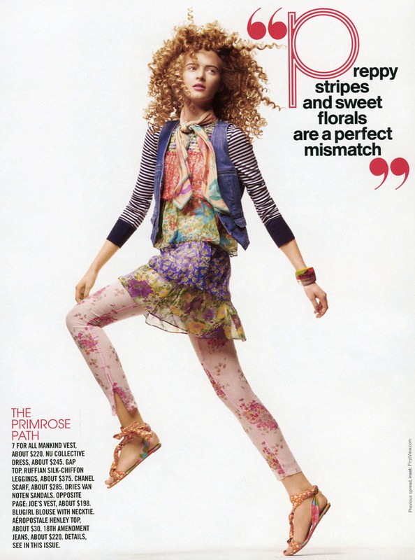 Fashion editorial shot of a young adult woman with curly red hair wearing a floral dress, leggings, and sandals in a studio setting.